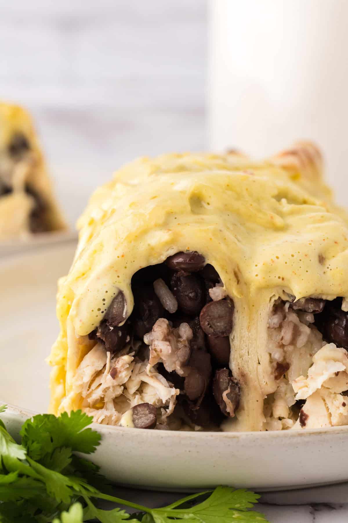 A close-up of a burrito cut open, showing rice, black beans, and shredded chicken inside, topped with a creamy yellow sauce. Fresh cilantro garnish is visible on the plate.
