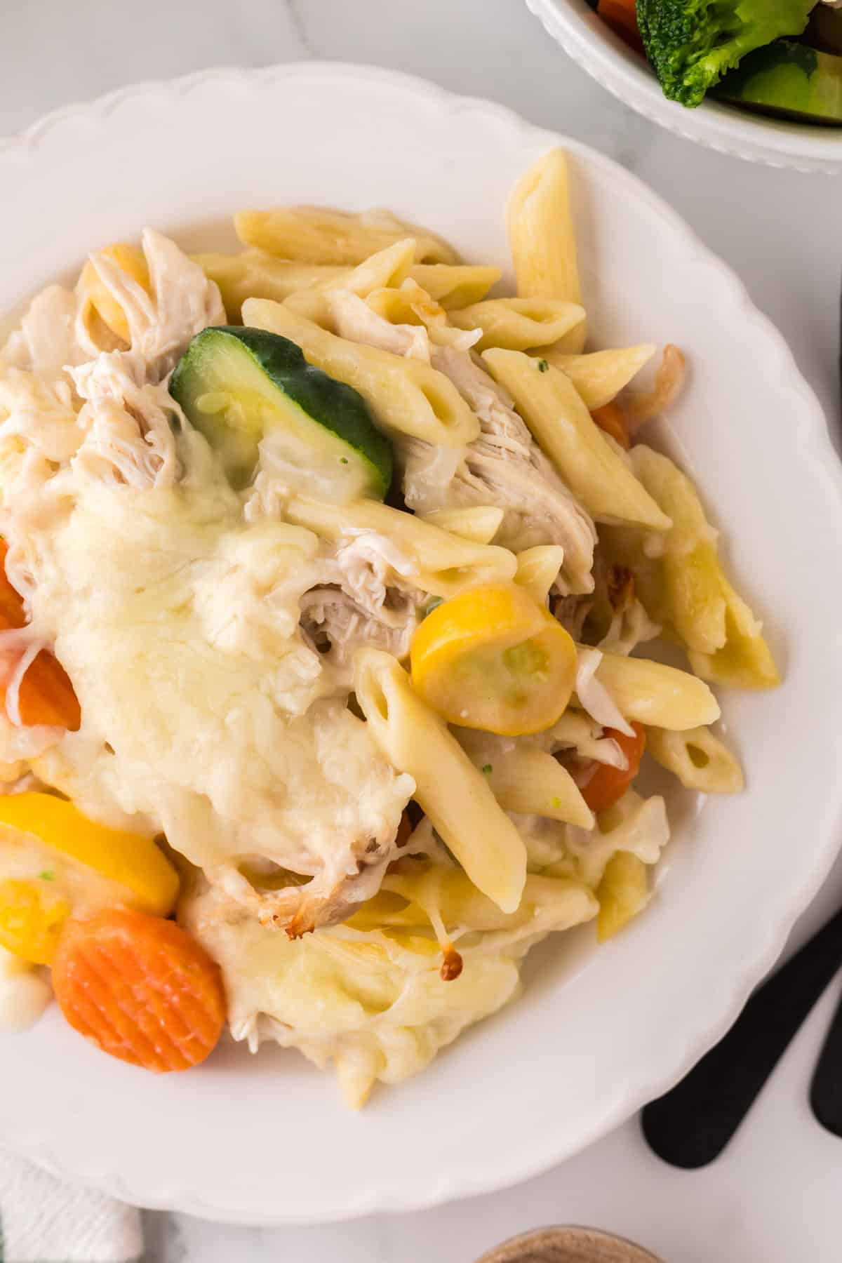 A white plate filled with chicken vegetable pasta casserole, topped with melted cheese, shredded chicken, zucchini slices, yellow squash, and carrots. A fork rests beside the plate on a white table.