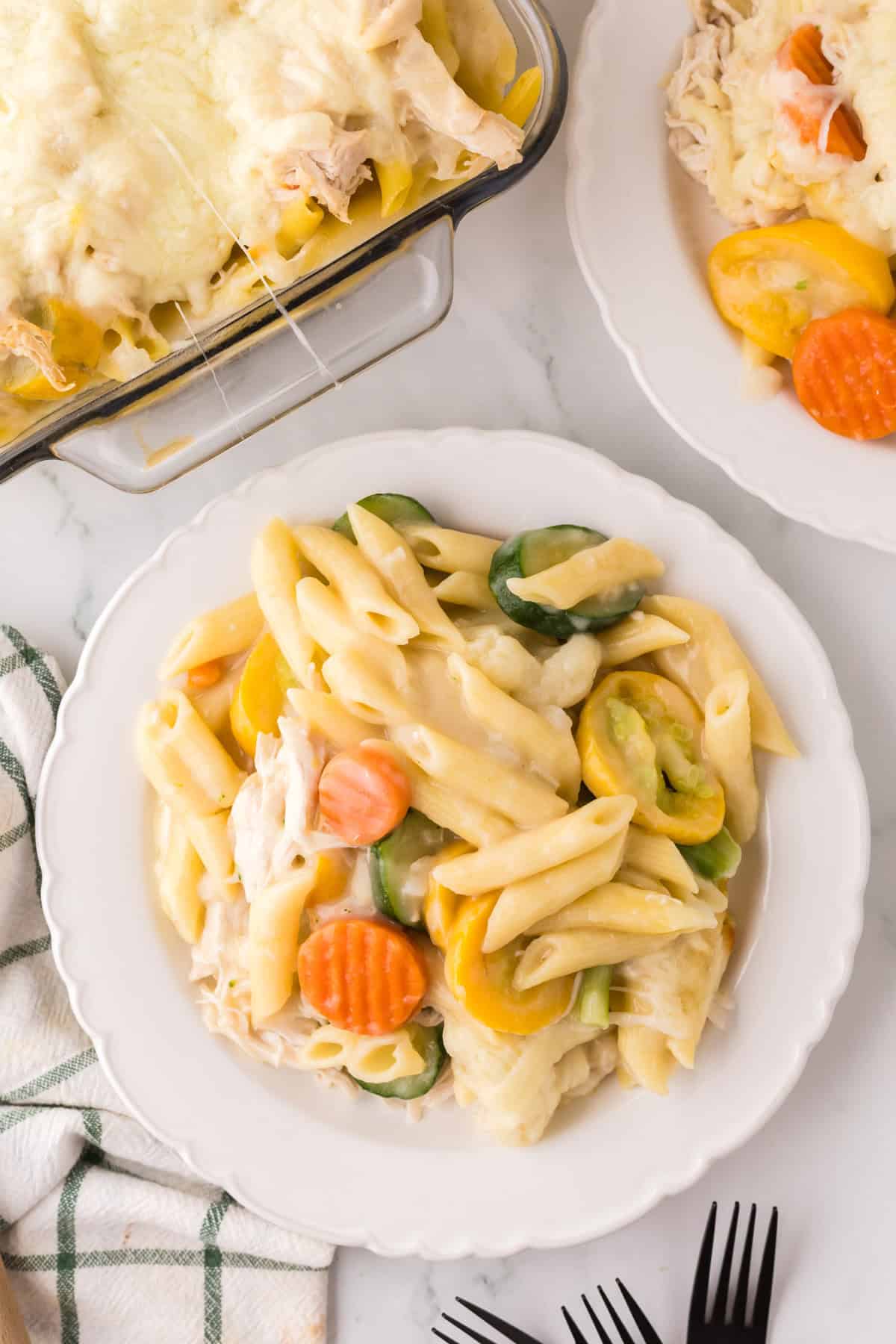 A white plate holds creamy chicken vegetable pasta mixed with sliced carrots, yellow squash, and zucchini, with a cheesy baked pasta dish and another plated serving in the background. A green-striped napkin and black forks are nearby.