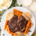 A plate of pot roast made in a slow cooker with cooked carrots and onion slices, garnished with herbs, sits next to a bowl of mashed potatoes and fresh rosemary and thyme on a marble surface. Gold utensils and a striped towel are nearby.