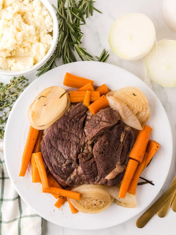 A plate of pot roast made in a slow cooker with cooked carrots and onion slices, garnished with herbs, sits next to a bowl of mashed potatoes and fresh rosemary and thyme on a marble surface. Gold utensils and a striped towel are nearby.