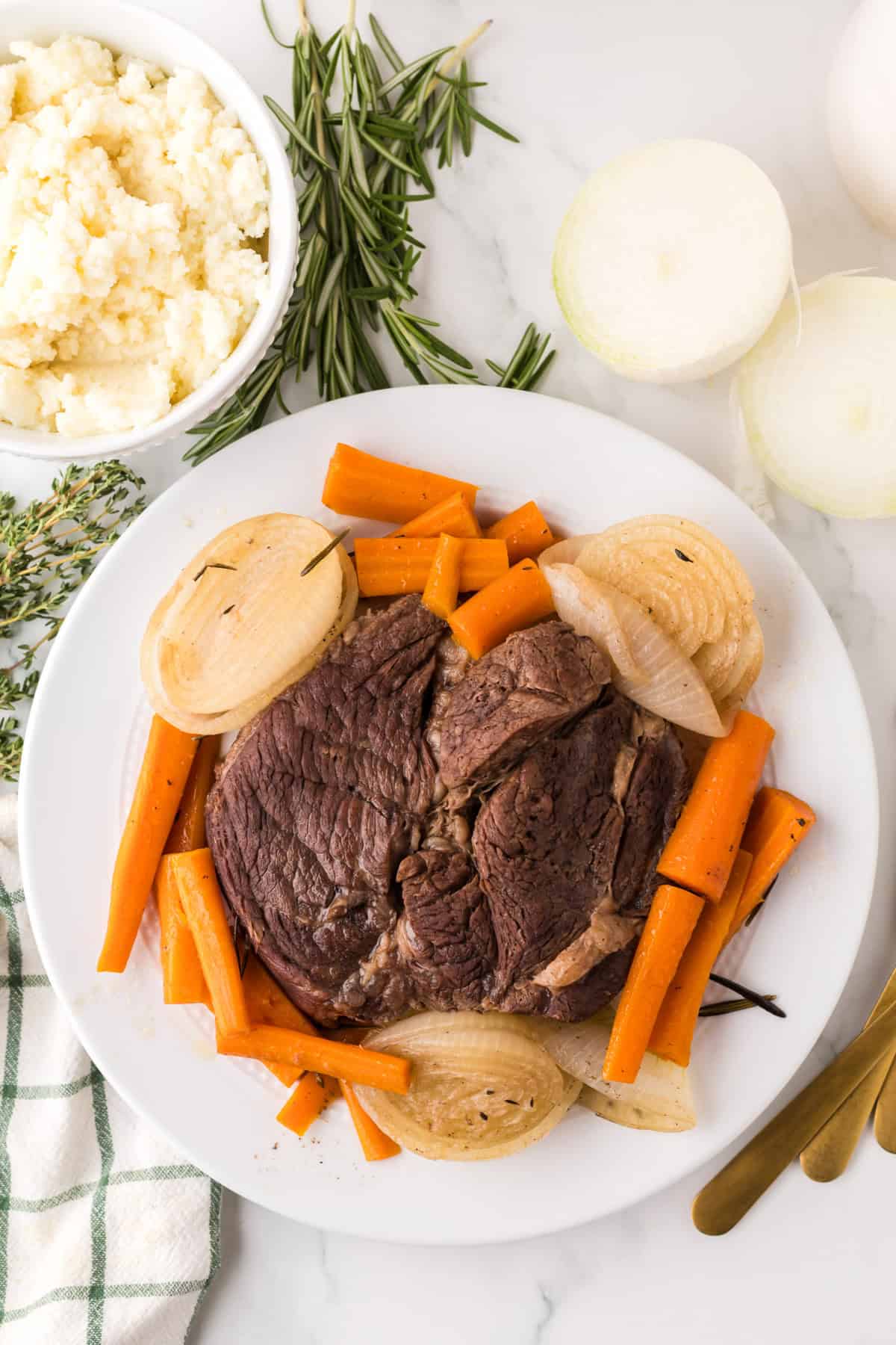 A plate of pot roast made in a slow cooker with cooked carrots and onion slices, garnished with herbs, sits next to a bowl of mashed potatoes and fresh rosemary and thyme on a marble surface. Gold utensils and a striped towel are nearby.
