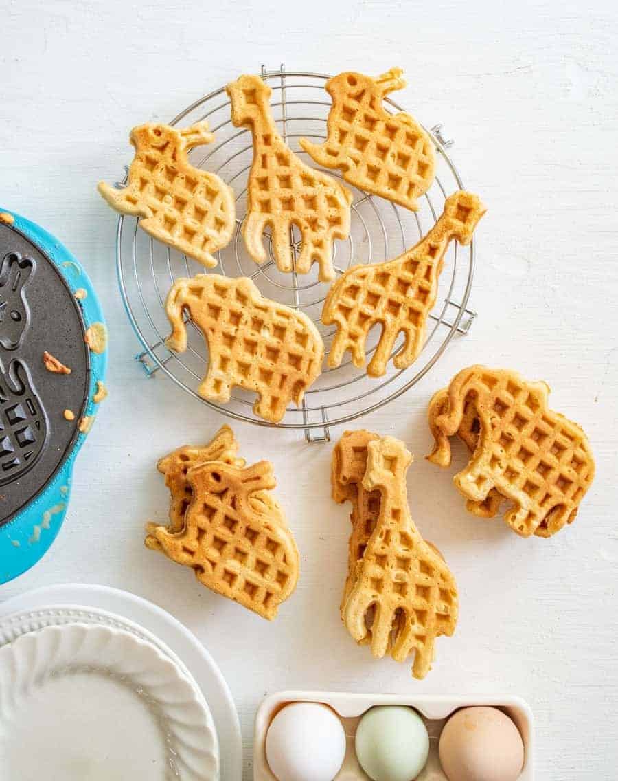 Animal-shaped waffles, including giraffes, elephants, and rhinos, are arranged on a cooling rack and white surface&mdash;perfect for adding fun to any easy weekly meal plan. Nearby sit a waffle maker, empty plates, and a carton with three eggs.