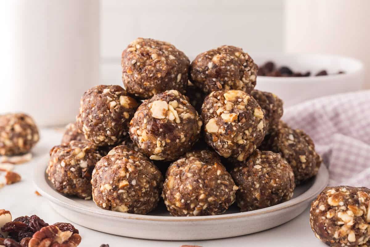 A plate stacked with round, brown energy bites made from chopped nuts and dried fruit&mdash;perfect for an easy weekly meal plan&mdash;with more bites and ingredients scattered around on a white surface.
