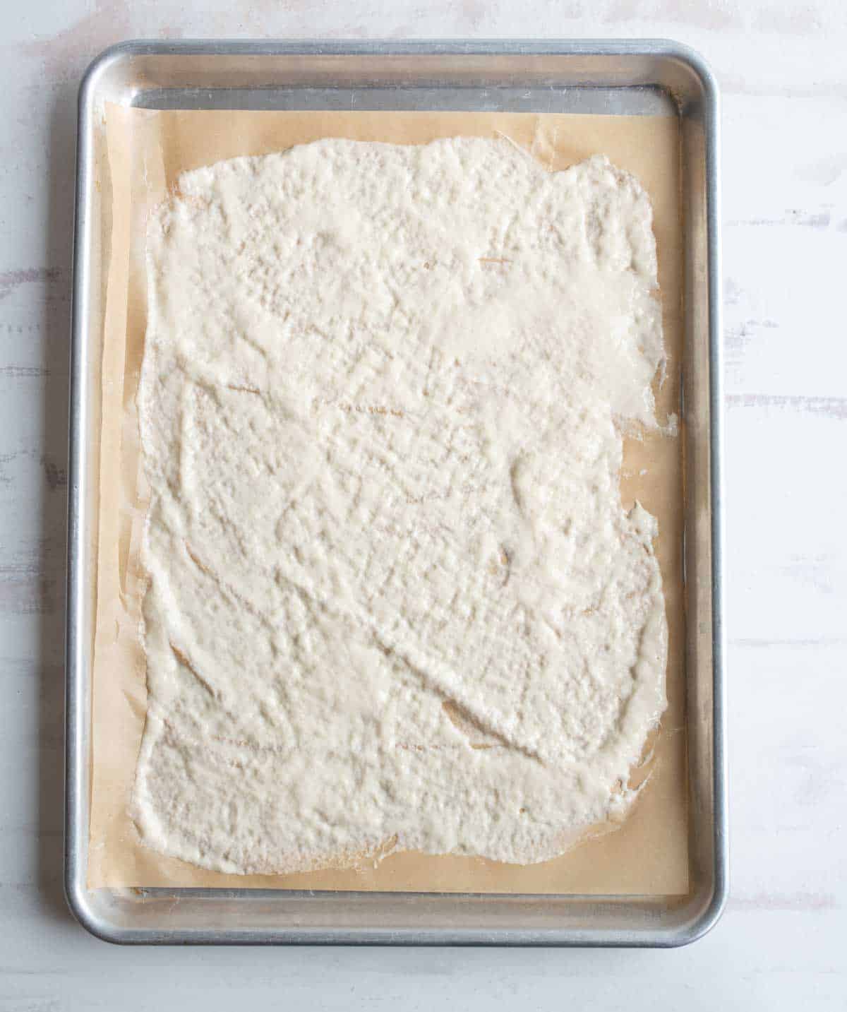 A baking sheet lined with parchment paper holds an unevenly spread layer of white, textured mixture, possibly dough or batter, ready to be baked.