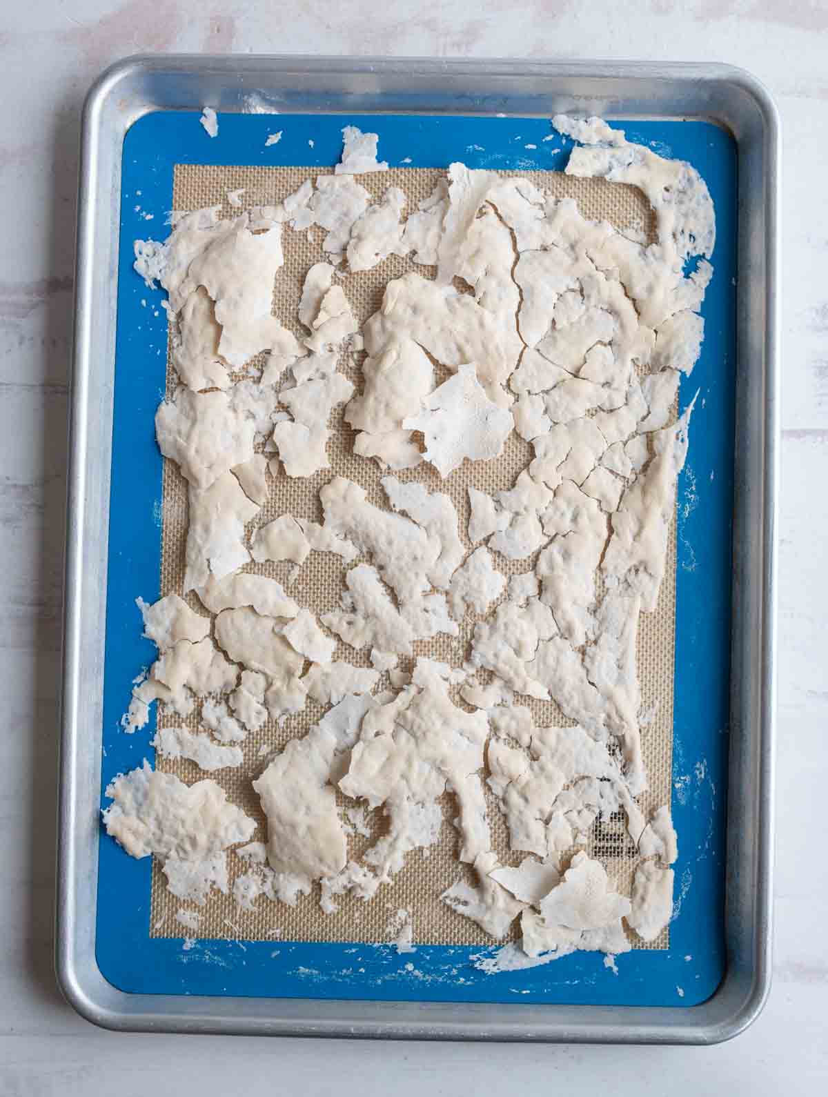 A baking sheet lined with a silicone mat holds unevenly spread, partially dried, torn pieces of dough on a light surface.