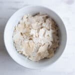 A white bowl filled with pale, irregular flakes of dried yeast on a light, textured surface showing how to dehydrate sourdough starter.