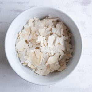 A white bowl filled with pale, irregular flakes of dried yeast on a light, textured surface showing how to dehydrate sourdough starter.