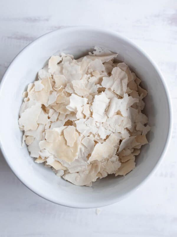 A white bowl filled with pale, irregular flakes of dried yeast on a light, textured surface showing how to dehydrate sourdough starter.