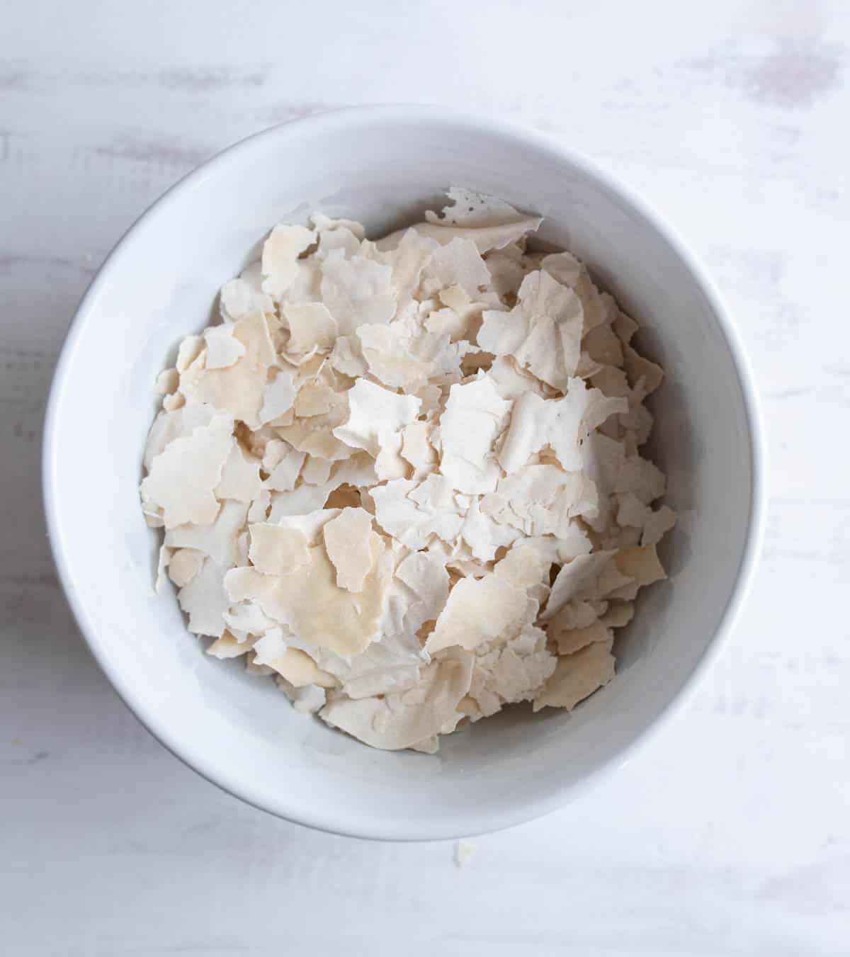 A white bowl filled with pale, irregular flakes of dried yeast on a light, textured surface showing how to dehydrate sourdough starter.