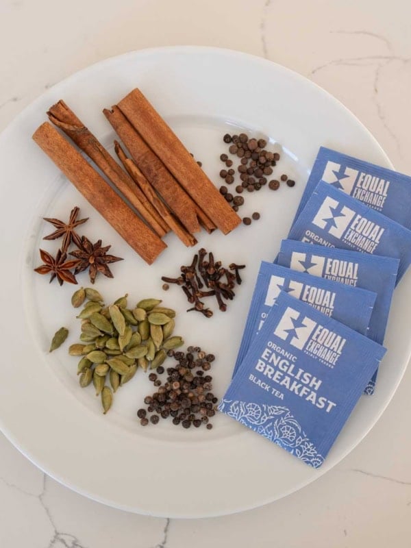 A white plate holds spices for a chai tea concentrate recipe—cinnamon sticks, cardamom pods, star anise, peppercorns, cloves—and four blue packets of Equal Exchange Organic English Breakfast black tea, all on a white marble surface.