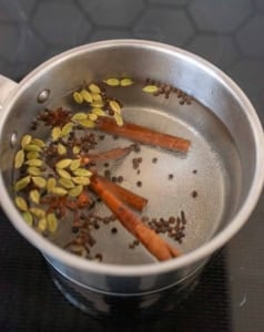 A silver pot on a stovetop contains water with cinnamon sticks, green cardamom pods, and whole cloves and star anise floating on the surface.
