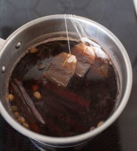 A saucepan on a stovetop contains dark liquid with tea bags, cinnamon sticks, and cardamom pods steeping inside.