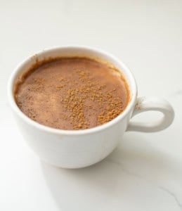 A white ceramic cup filled with frothy hot chocolate, topped with a light dusting of cocoa powder, sits on a white surface.