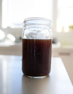 A glass mason jar filled with dark brown liquid sits on a white countertop, with a bright, sunlit kitchen blurred in the background.