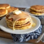 A stack of cornmeal pancakes topped with melting butter and syrup on a white plate. There are more pancakes in the background on a cooling rack and a separate plate. Forks are placed beside the dish on a striped cloth.