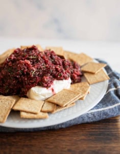 A plate topped with a mound of cranberry salsa on cream cheese, surrounded by square crackers. The setting is on a wooden table with a blue cloth underneath the plate.