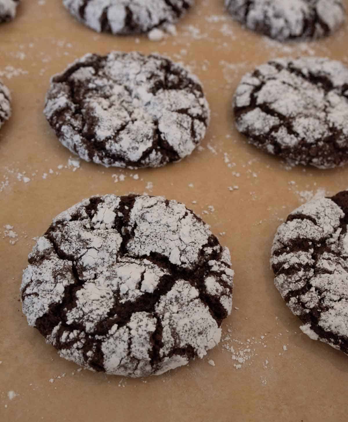 Chocolate crinkle cookies dusted with powdered sugar are displayed on a parchment-lined baking sheet. The cookies have a cracked surface, revealing the chocolate underneath.