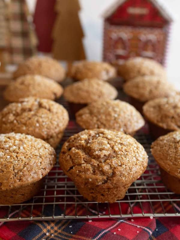 A batch of freshly baked gingerbread muffins topped with sugar crystals resting on a cooling rack. In the background, there are decorative, holiday-themed wooden trees and a red, house-shaped ornament.