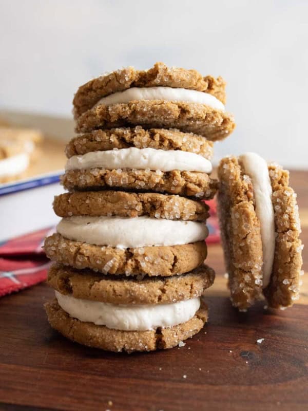 A stack of gingerbread sandwich cookies with white cream filling is arranged on a wooden surface. One cookie is leaning against the stack. A red plaid cloth is partially visible in the background.
