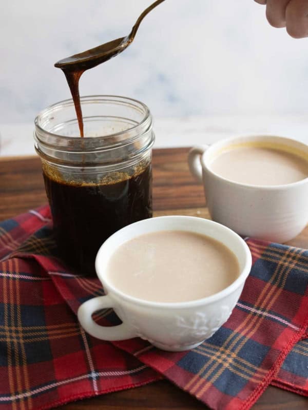 A jar of dark gingerbread syrup with a spoon above it, next to two cups of coffee with cream. The setup is on a wooden surface with a red plaid cloth underneath.