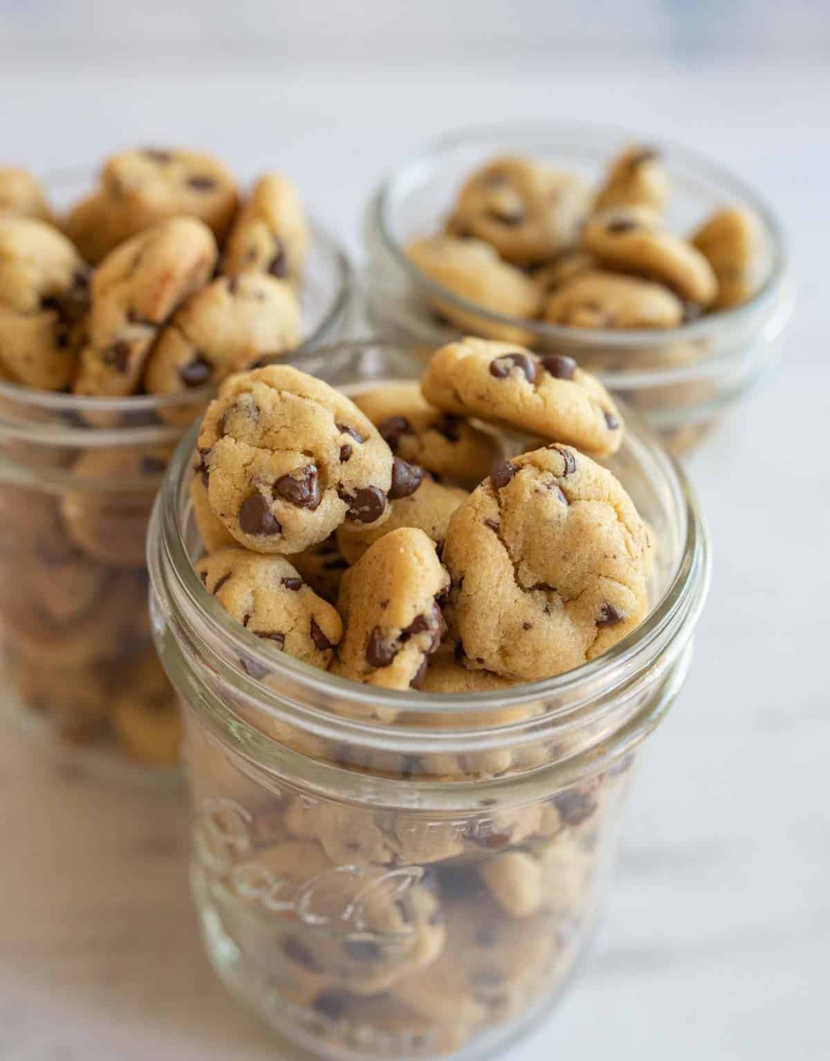 Three glass jars filled with small chocolate chip cookies are placed on a white marble surface. The cookies are golden-brown and plentiful, with visible chocolate chips, creating a tempting display.