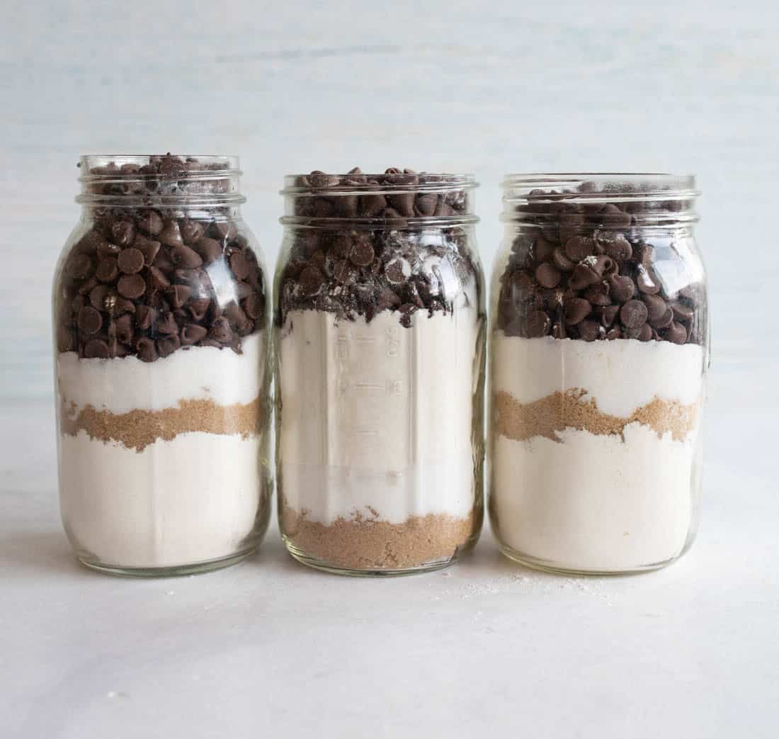 Three glass jars filled with layers of baking ingredients: chocolate chips, flour, white sugar, and brown sugar, standing on a light surface with a neutral background.
