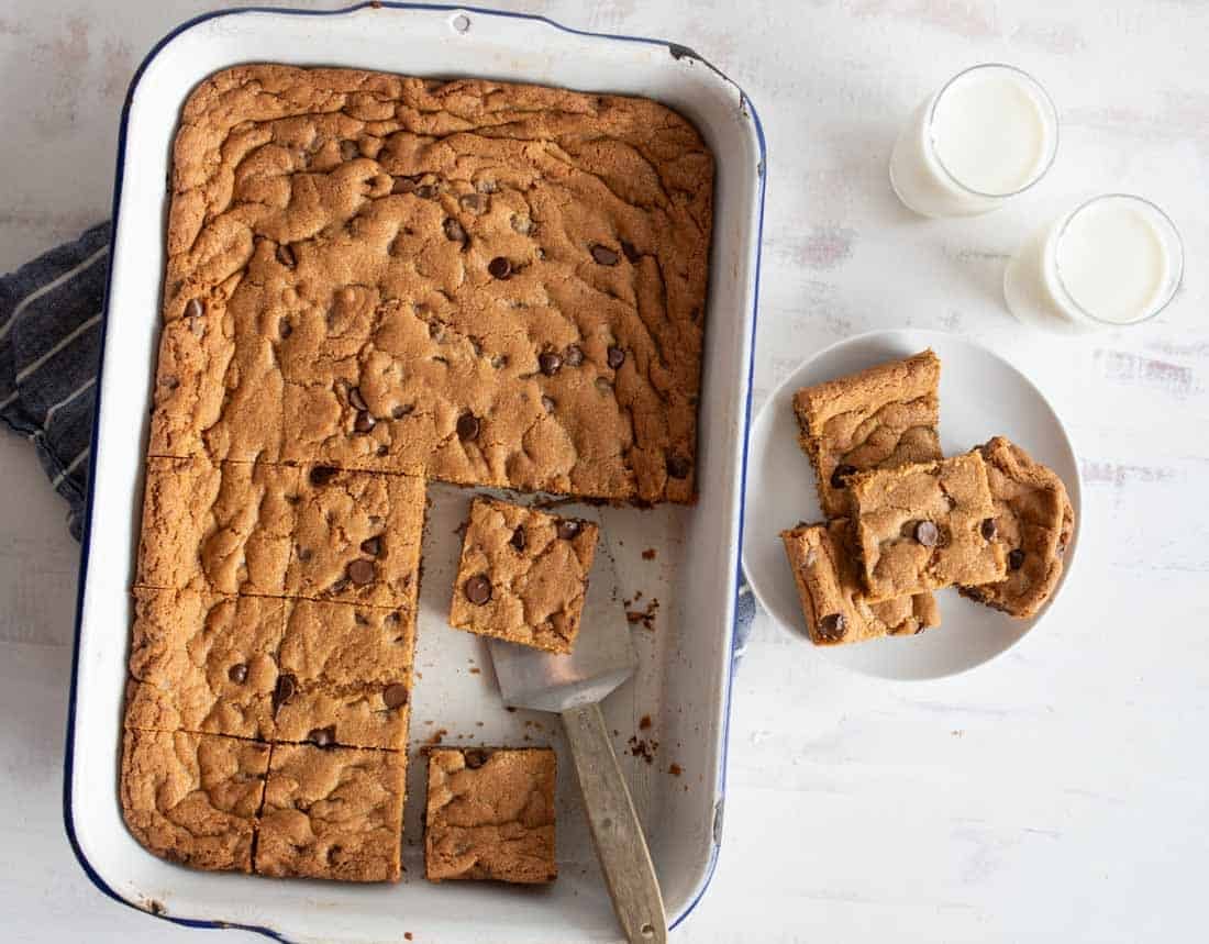 A baking dish with sliced chocolate chip cookie bars. Several bars are served on a white plate beside the dish. Two glasses of milk sit nearby. A spatula rests in the baking dish. The surface is light and textured.