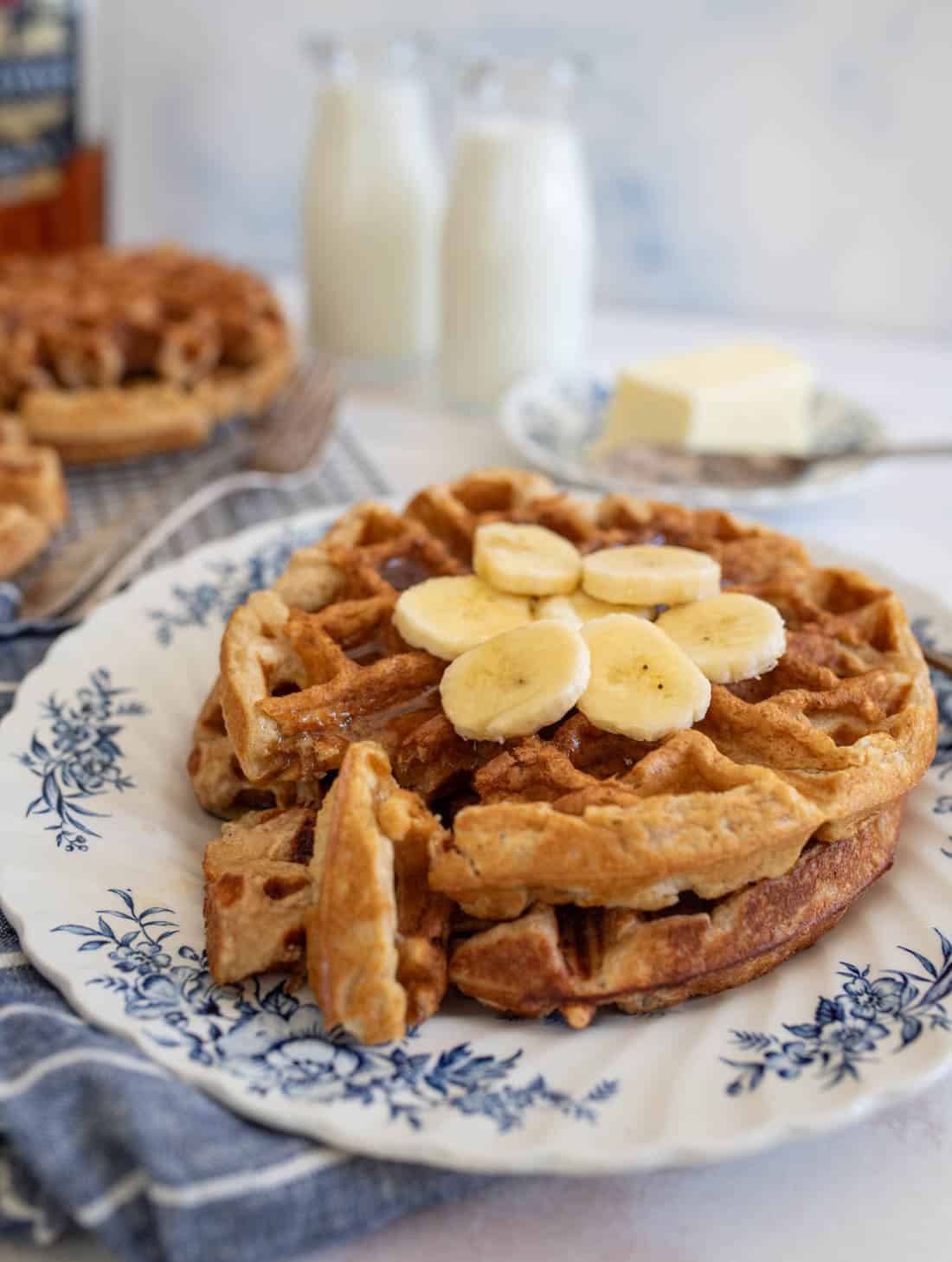A stack of waffles topped with banana slices on a floral-patterned plate. In the background, there are two bottles of milk and a block of butter on a small dish. Another waffle is on a cooling rack. A blue and white cloth is partially visible.