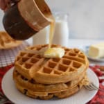 A hand pours syrup from a rustic cup onto a stack of waffles topped with butter on a white plate. A red cloth and a bottle of milk are in the background, along with a pat of butter and a fork.
