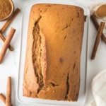 Freshly baked gingerbread loaf rests on a white cutting board, surrounded by cinnamon sticks and a small bowl of cocoa powder. A cup of dark liquid and a white cloth are nearby, set on a white marble surface.