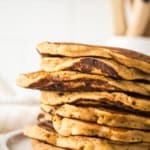 A stack of golden-brown gingerbread pancakes on a white plate, with a blurred background of wooden kitchen utensils in a holder. The pancakes have a fluffy texture and a slightly crisp edge.