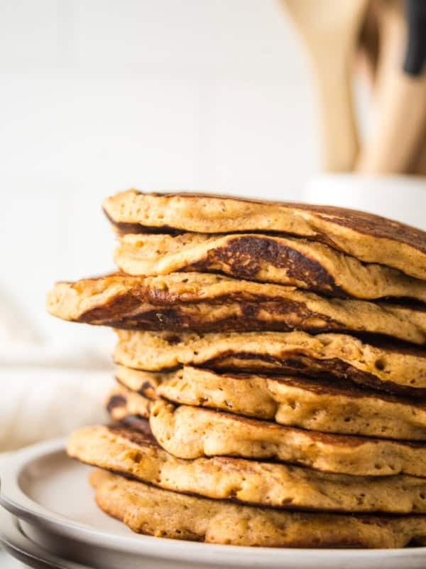 A stack of golden-brown gingerbread pancakes on a white plate, with a blurred background of wooden kitchen utensils in a holder. The pancakes have a fluffy texture and a slightly crisp edge.