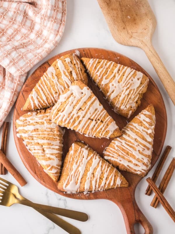 Triangle-shaped gingerbread scones with white icing drizzle sit on a round wooden board. A wooden spatula, checkered cloth, cinnamon sticks, a glass, and gold utensils surround the board on a marble surface.