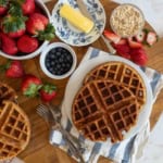 A wooden board with fresh strawberries, blueberries, oats, butter, and waffles on a plate. Forks rest beside the plate, and a striped napkin enhances the presentation.