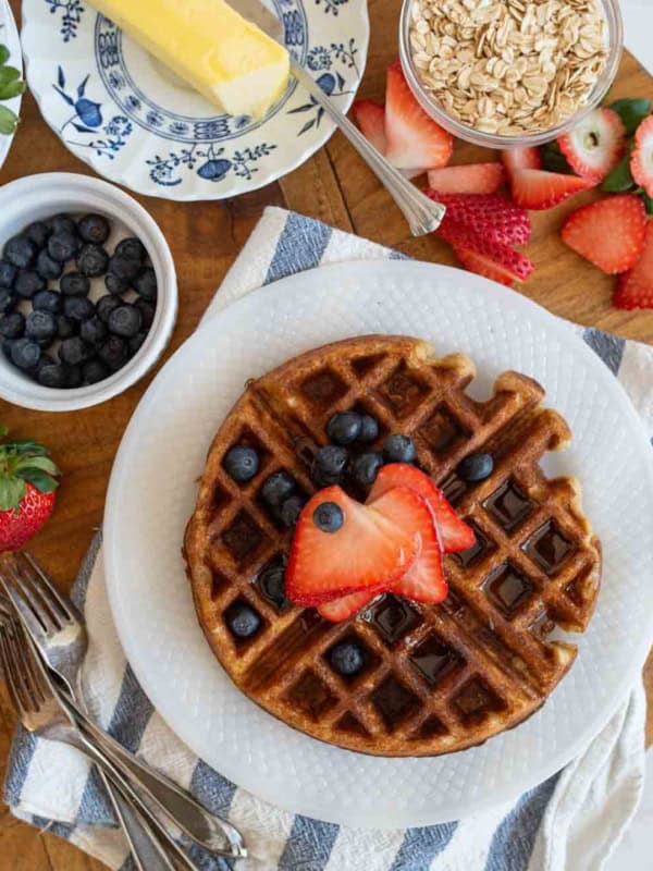 Oatmeal waffles topped with sliced strawberries and blueberries sits on a white plate. Nearby, theres a dish of blueberries, a butter dish with butter, a bowl of oats, sliced strawberries, and silverware on a striped napkin, all on a wooden table.