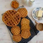 A cooling rack holds various sized golden-brown waffles on a checkered cloth. Nearby are jars of milk, a dish with butter, a small bowl of orange spread, and a mug of coffee, all arranged on a white surface.