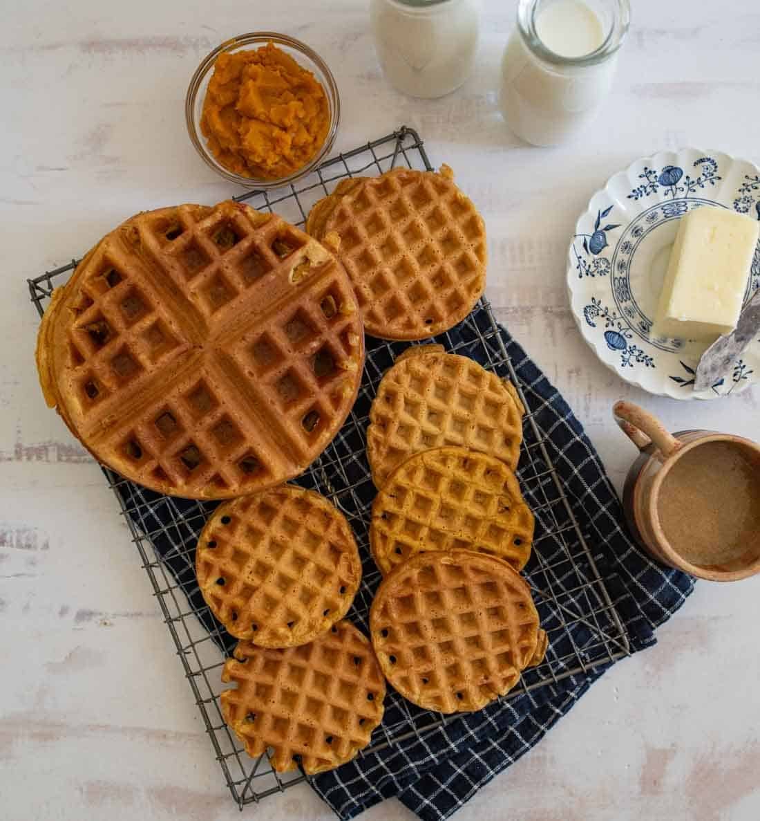A cooling rack holds various sized golden-brown waffles on a checkered cloth. Nearby are jars of milk, a dish with butter, a small bowl of orange spread, and a mug of coffee, all arranged on a white surface.