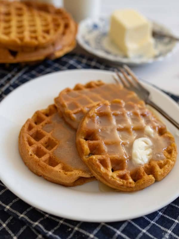 Two pumpkin waffles on a white plate, drizzled with syrup and topped with butter. A fork rests nearby. In the background, a stack of waffles and a dish with a butter block sit on a checkered cloth.