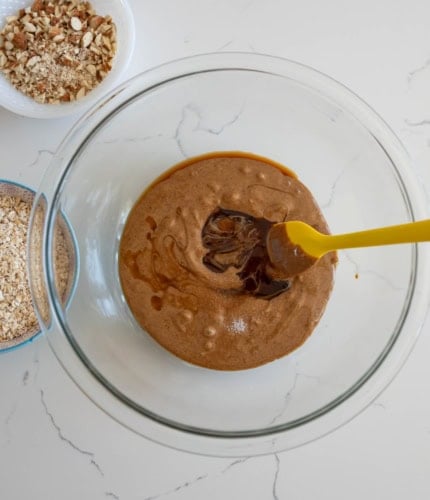 A glass bowl holds a smooth, brown mixture being stirred with a yellow spatula. Nearby, bowls with chopped nuts and oats are visible on a white, marbled surface.