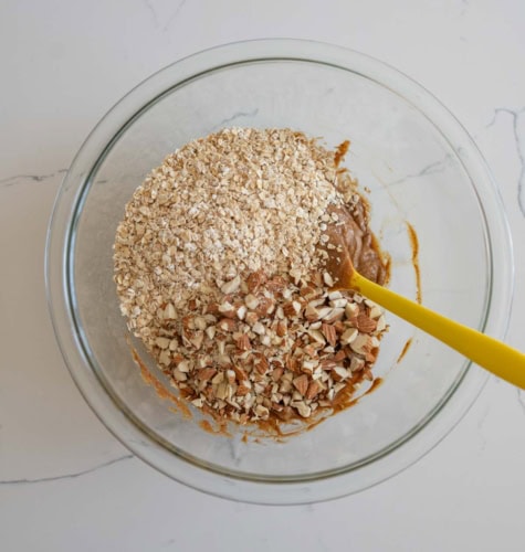 A glass bowl contains a mixture of oats, chopped almonds, and a brown batter, with a yellow spatula partially submerged in the ingredients. The bowl is placed on a white marble surface.