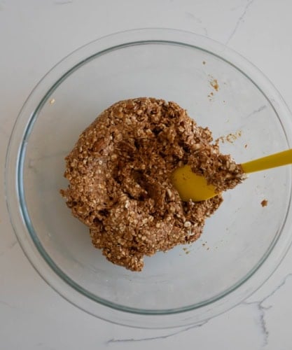 A glass bowl containing a mixture of oats and dough-like substance, being stirred with a yellow spatula. The bowl is placed on a white marble countertop.