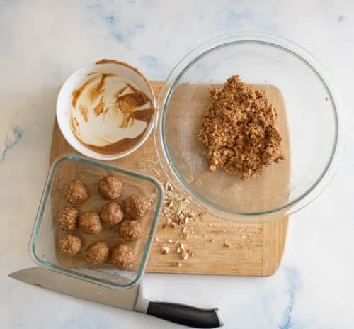 A cutting board with peanut butter protein balls, a glass bowl of dough, an empty bowl with peanut butter remnants, and a large knife. Some nuts are scattered on the board, all on a light marble surface.