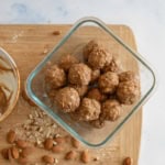 Glass container holding energy bites with oats and nuts, placed on a wooden cutting board sprinkled with crushed almonds. A used mixing bowl with residue is partially visible to the left.