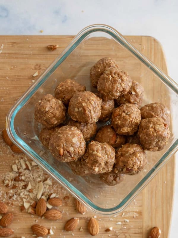 Glass container holding energy bites with oats and nuts, placed on a wooden cutting board sprinkled with crushed almonds. A used mixing bowl with residue is partially visible to the left.