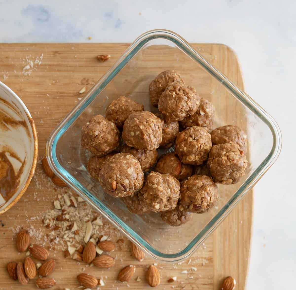 Glass container holding energy bites with oats and nuts, placed on a wooden cutting board sprinkled with crushed almonds. A used mixing bowl with residue is partially visible to the left.