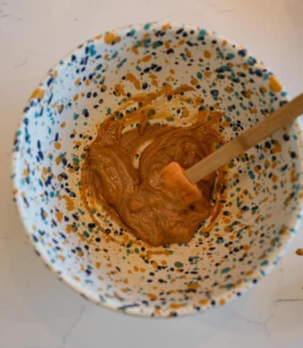 A ceramic bowl with a colorful speckled pattern holds a creamy brown mixture. An orange spatula with a wooden handle is resting in the mixture, which appears to be partially blended. The bowl is on a white surface.