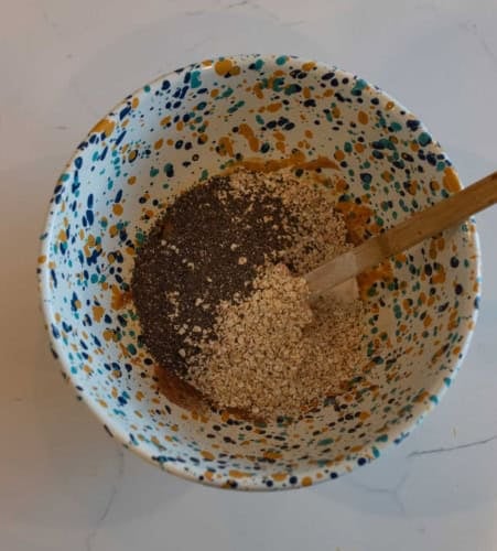 A speckled bowl contains a mixture of oats and chia seeds, with a wooden spoon resting on the side, on a white marble surface.