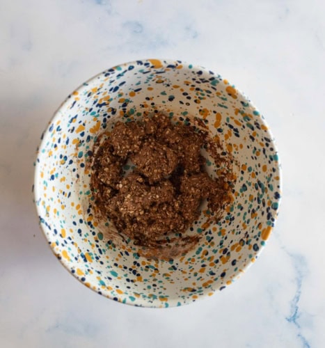 A speckled bowl containing a mixture of brown ingredients, likely a type of oatmeal or batter, is placed on a light marble surface. The bowl has colorful spots in blue, yellow, and green.