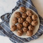 A white plate with several round, brown chocolate peanut butter protein balls topped with oats. The plate is placed on a crumpled, blue and white striped cloth on a marble surface.